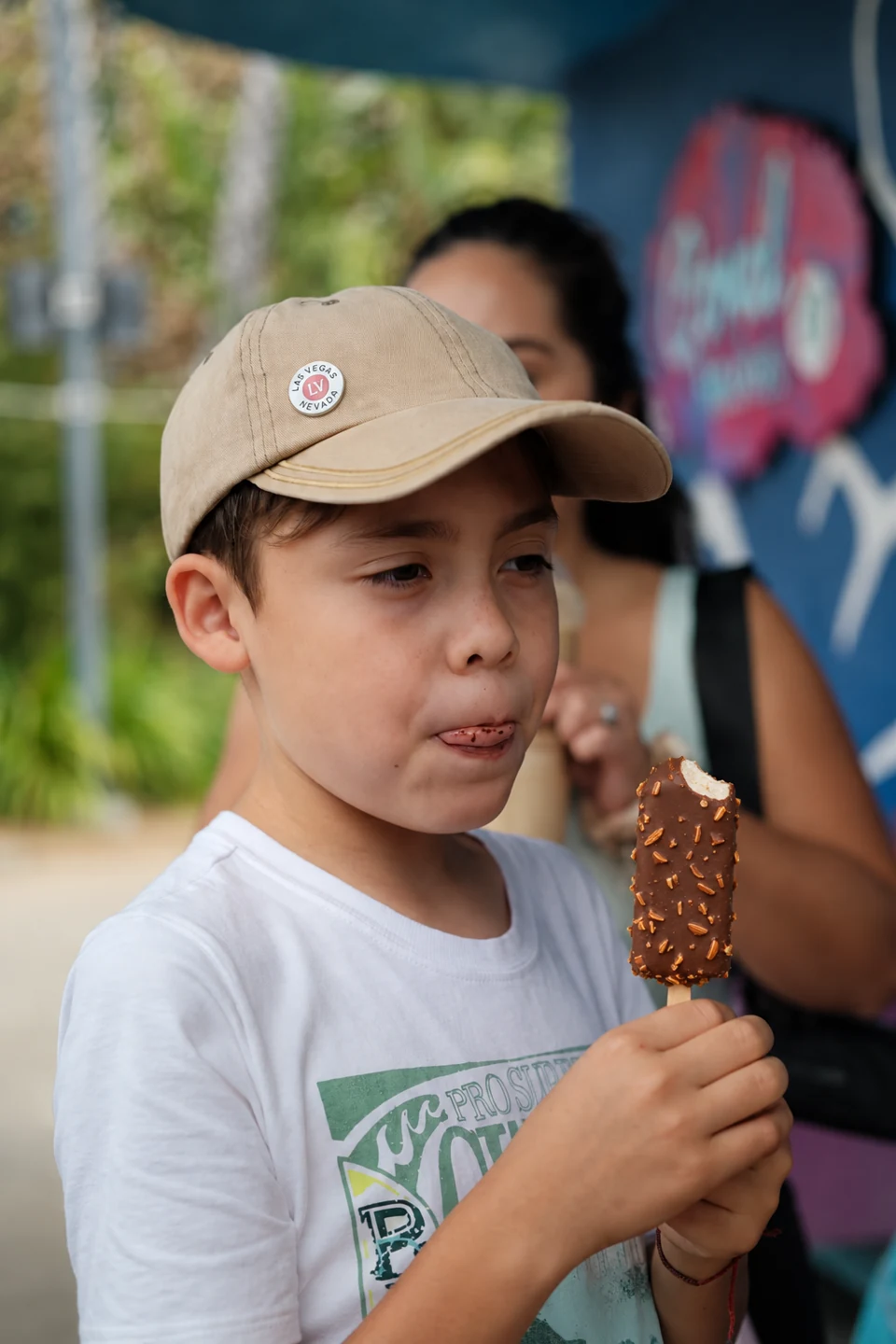 A child in a beige cap enjoying a chocolate almond ice cream at an outdoor kiosk